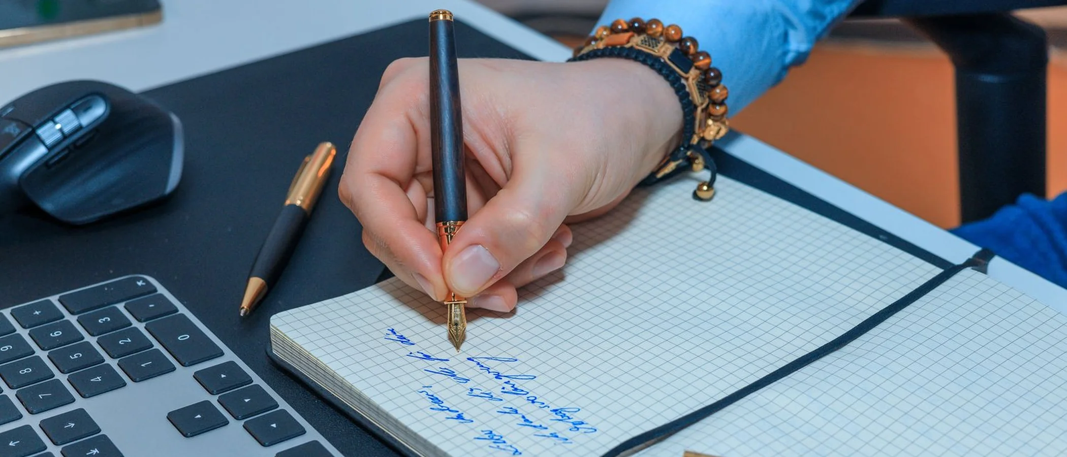 A hand writing in a grid notebook with an ebony and gold fountain pen at a desk