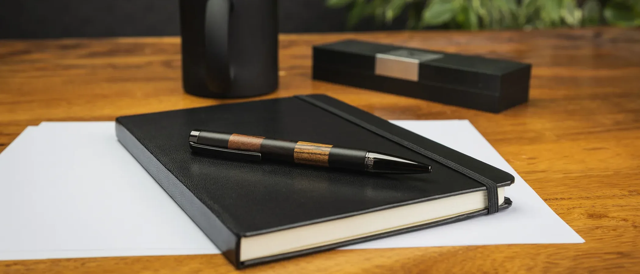 A wood-accent pen resting on a black notebook on a wooden desk, with a mug and a gift box