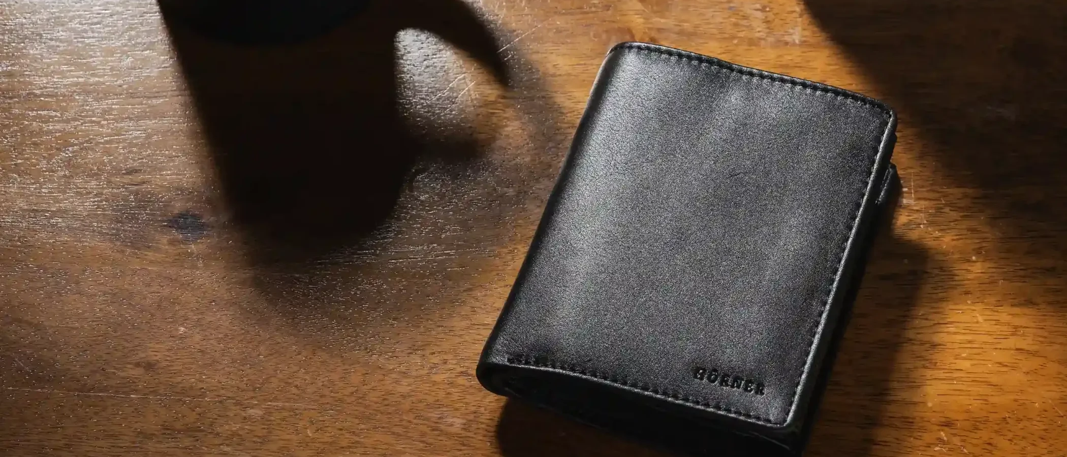 A black leather Hörner wallet on a dark wood desk beside a coffee cup