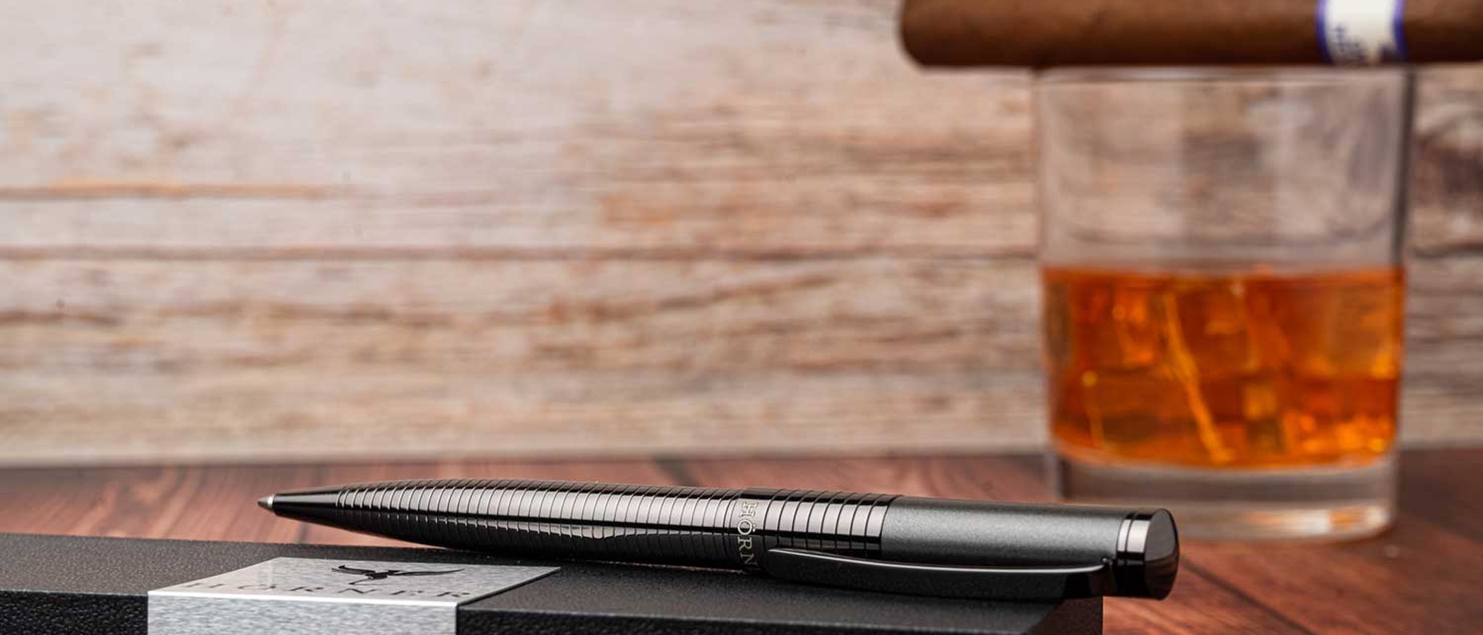 A gunmetal Hörner ballpoint pen resting on its gift box beside a whiskey and cigar